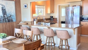A modern kitchen with wooden cabinets, a stainless steel refrigerator, and a dining area with a wooden table and chairs. - Best Location! Best View! - EXCLUSIVELY ON VRBO (Cayucos)