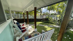 A covered patio with white chairs and a bench, stone flooring, and a view of a garden with grass and trees. - Footsteps to Hawaii's Best Beach  (Kailua)