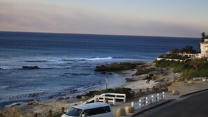 Una carretera costera con una camioneta blanca estacionada al lado, que conduce a una playa con terreno rocoso y algunas personas caminando.