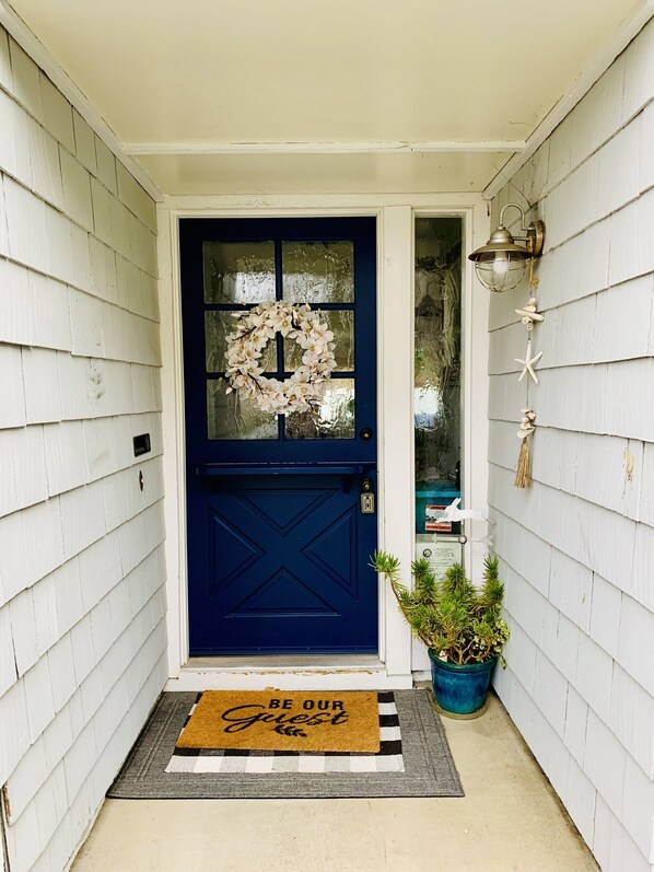 A blue front door with a wreath, a doormat with "BE OUR GUEST" text, and a potted plant beside the door.