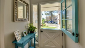 Un couloir avec une porte donnant sur l’extérieur, une console bleue ornée de cadres et un miroir accroché au mur.