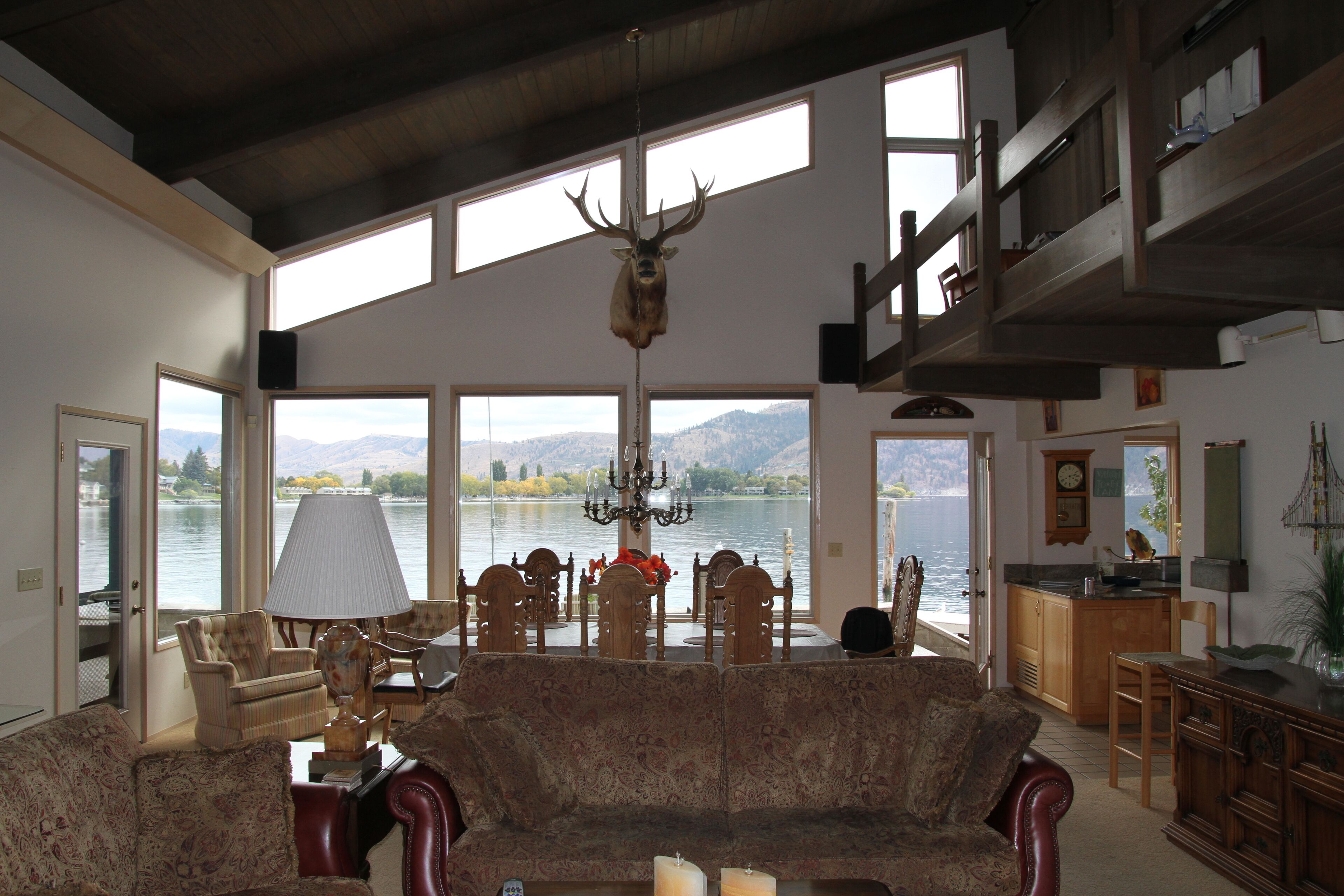 Un vaste salon doté d’une grande fenêtre donnant sur un lac, d’un lustre et d’une tête de cerf fixée au mur.