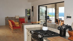 A modern kitchen with a gas stove, a dining area with a table and chairs, and a view of the ocean through a glass door. - Architecturally Designed Villa in Paradise, Quiet Beach & Blue Water Views (Turtle Beach)