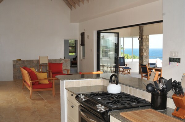 A modern kitchen with a gas stove, a dining area with a table and chairs, and a view of the ocean through a glass door. - Architecturally Designed Villa in Paradise, Quiet Beach & Blue Water Views (Turtle Beach)