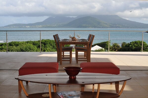 A dining area with a table and chairs, a red cushioned bench, and a view of the sea and mountains.