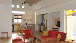 A spacious living area with a kitchen in the background, wooden furniture, and a large window providing natural light. - Architecturally Designed Villa in Paradise, Quiet Beach & Blue Water Views (Turtle Beach)