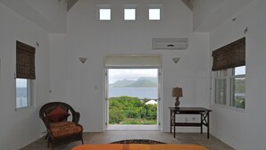 A bedroom with a bed, a chair, a table, and a view of the sea and mountains. - Architecturally Designed Villa in Paradise, Quiet Beach & Blue Water Views (Turtle Beach)
