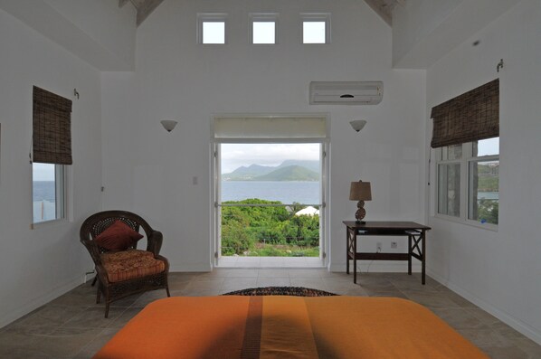 A bedroom with a bed, a chair, a table, and a view of the sea and mountains. - Architecturally Designed Villa in Paradise, Quiet Beach & Blue Water Views (Turtle Beach)