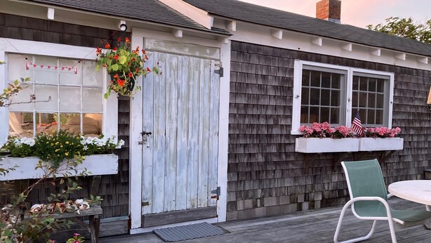 A wooden deck with a white door, a window with flowers, and a green chair. - The Quintessential Traditional Nantucket Vacation! (Nantucket)