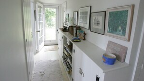 A narrow hallway with white walls, a white door, and a window leading to a bright outdoor area. - The Quintessential Traditional Nantucket Vacation! (Nantucket)