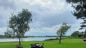 Een houten terras met twee stoelen en een tafel, met uitzicht op een gazon met water en een voertuig in de verte.