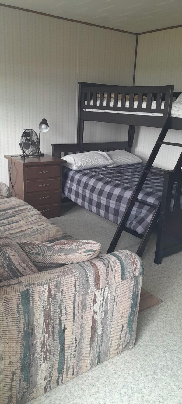 Uma cama beliche com colcha xadrez, uma mesinha de cabeceira de madeira com abajur e um sofá estampado.
