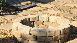 Un pozo de fuego de piedra en un jardín con una terraza de madera y un edificio blanco al fondo.