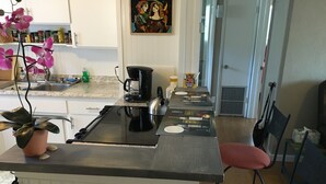 Una cocina con una isla central, cafetera y lavamanos. Hay dos taburetes rojos y una planta en maceta sobre la isla.