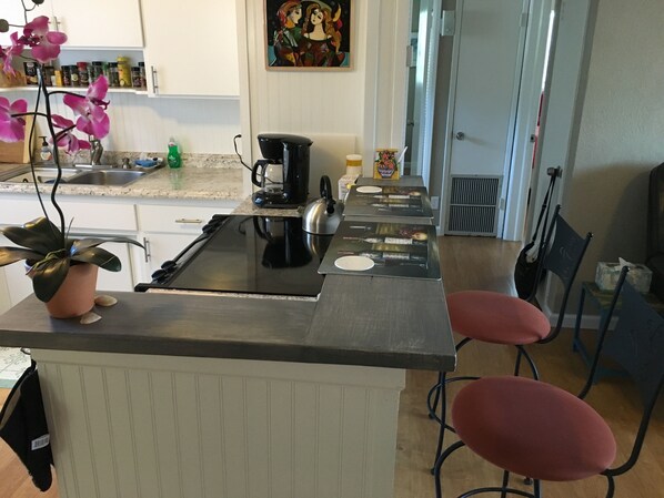 Una cocina con una isla central, cafetera y lavamanos. Hay dos taburetes rojos y una planta en maceta sobre la isla.