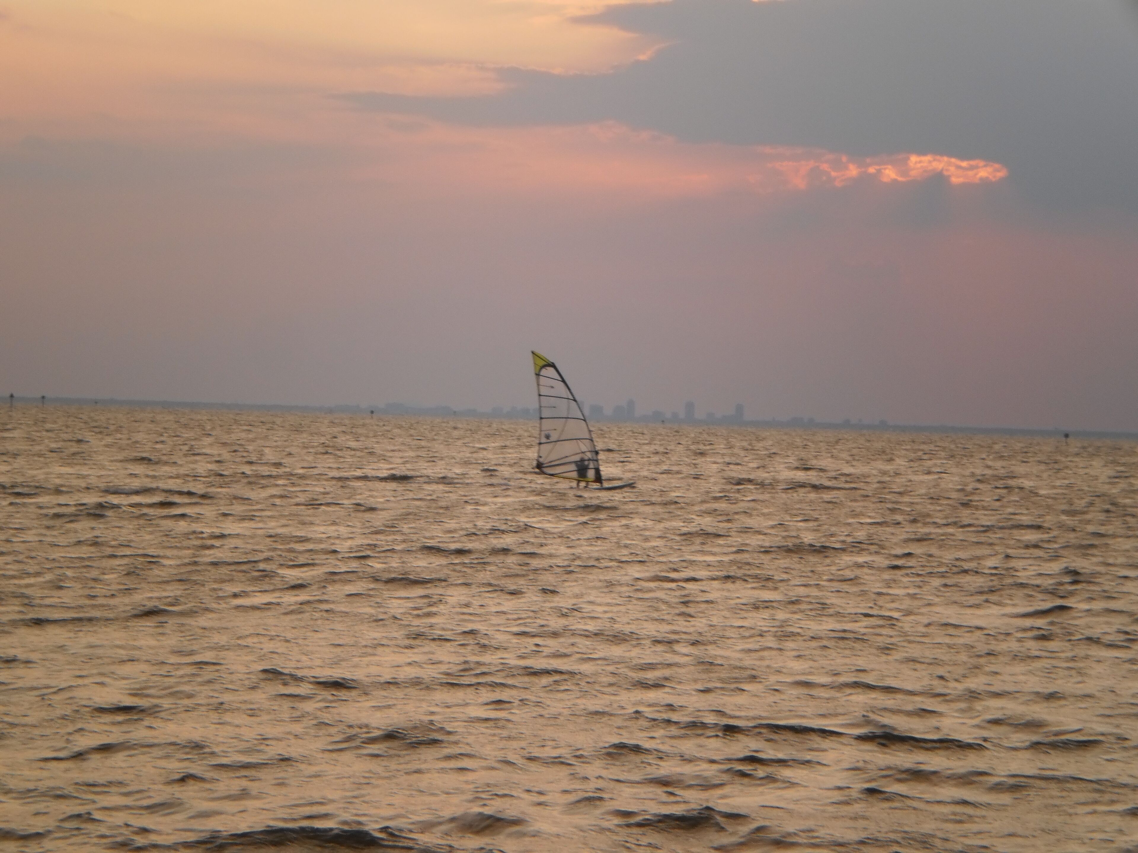 Una persona practicando windsurf en un mar tranquilo, con un horizonte urbano de fondo.