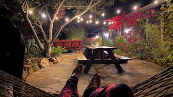 A patio area with a wooden table and benches, surrounded by plants and a string of lights.