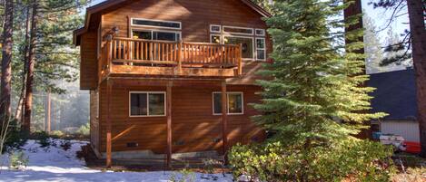 A two-story wooden cabin with a balcony, surrounded by tall pine trees and snow on the ground.