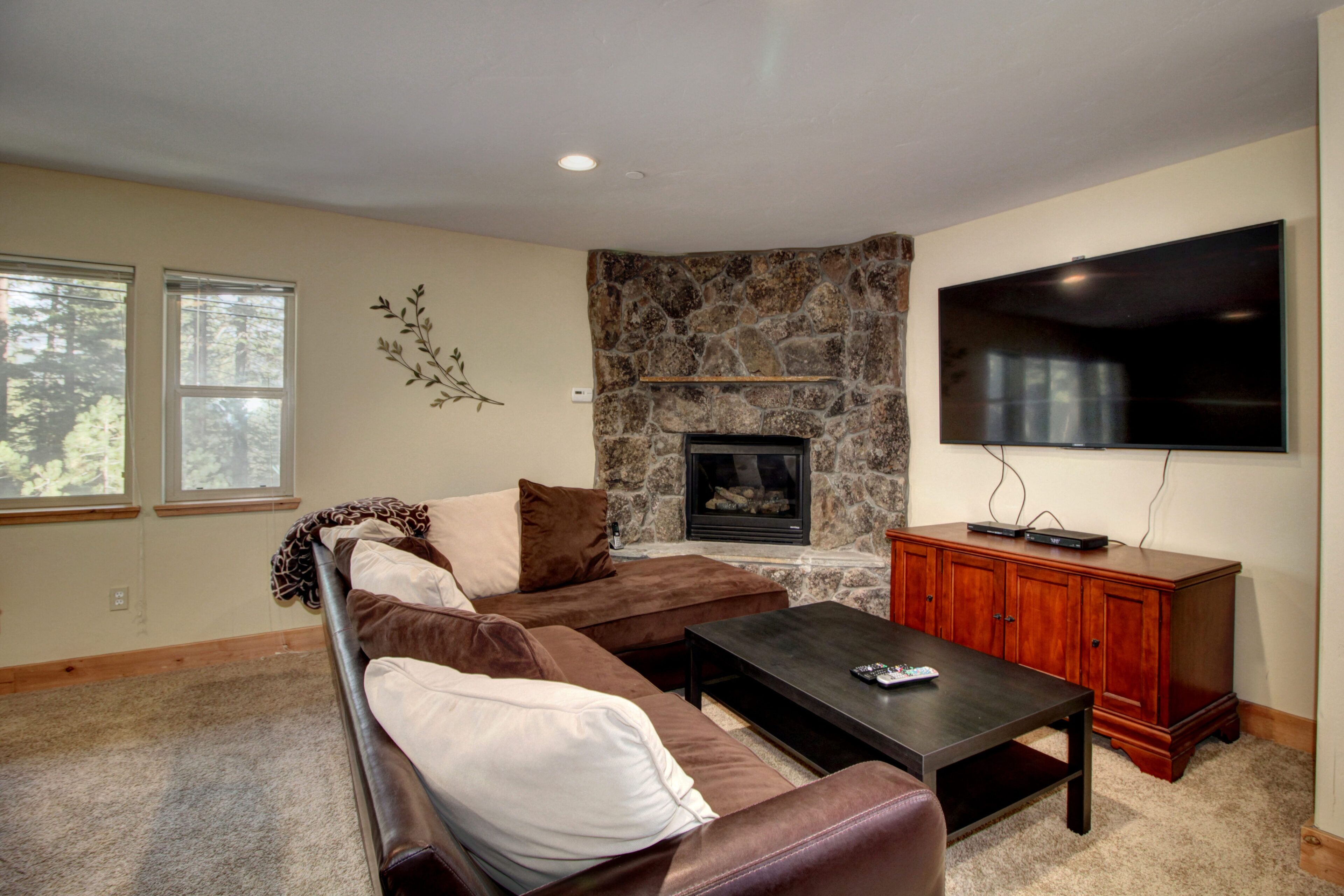 A living room with a stone fireplace, a large flat-screen TV, a brown leather sofa, and a wooden cabinet.
