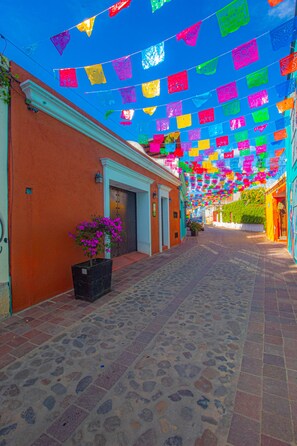Une rue étroite, ornée de guirlandes de drapeaux colorés suspendues au-dessus, avec un pavé de pierre et un bâtiment dont la porte et la fenêtre se trouvent sur le côté gauche.