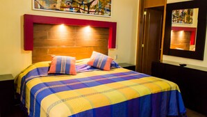 Ein Hotelzimmer mit einem großen Bett, einem Holz-Kopfteil, einem Gemälde darüber, einem Spiegel an der Wand und einer Tür zur rechten Seite.