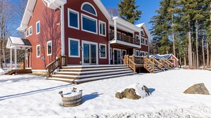 A large, two-story house with a red exterior and white trim, featuring a front porch and a staircase leading to a second-floor balcony. - Luxury Mountain Home with Hot Tub, Sauna & Views — Walk to Mountain Top Inn (Chittenden)