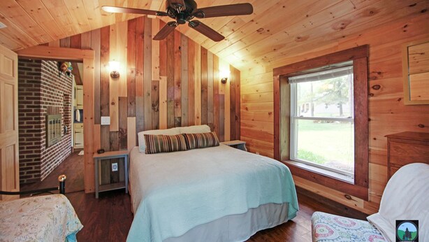Ein Schlafzimmer einer Holzhütte mit Bett, Fenster, Deckenventilator und Backsteinwand.