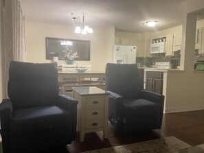 Un salon avec deux fauteuils sombres, une petite table basse et un coin repas comprenant une table et des chaises. On aperçoit une cuisine à l’arrière-plan, avec des placards et des appareils électroménagers.