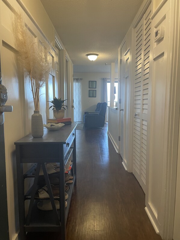 A hallway with a wooden floor, a table with a vase and a plant, a chair, and a door with a thermostat. - Weeks in September still available! Book now for a great vacation. (Myrtle Beach)