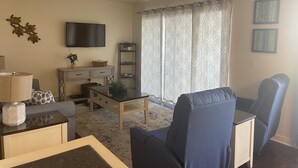 A living room with a sofa, a coffee table, a TV, a bookshelf, and a window with curtains. - Weeks in September still available! Book now for a great vacation. (Myrtle Beach)