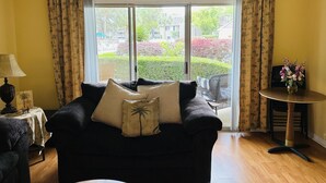Een woonkamer met een bank, een bijzettafeltje met een lamp en uitzicht op een tuin door een groot raam.