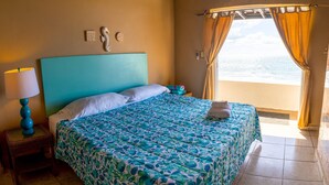 Un dormitorio con cama, mesita de noche, lámpara y vista al océano a través de la ventana.