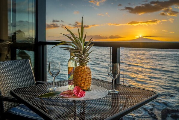 Una mesa con una piña, una botella de licor y vasos, frente a una vista al atardecer desde un balcón.