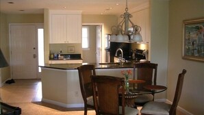 A compact kitchen with white cabinets, a central island, and a dining area with wooden chairs and a glass table. - PANORAMIC GOLF LAGOON VIEW,WALK TO BEACH, BEACH LOCKER,FROM OWNER,NO HIDDEN FEES (Hilton Head Island)