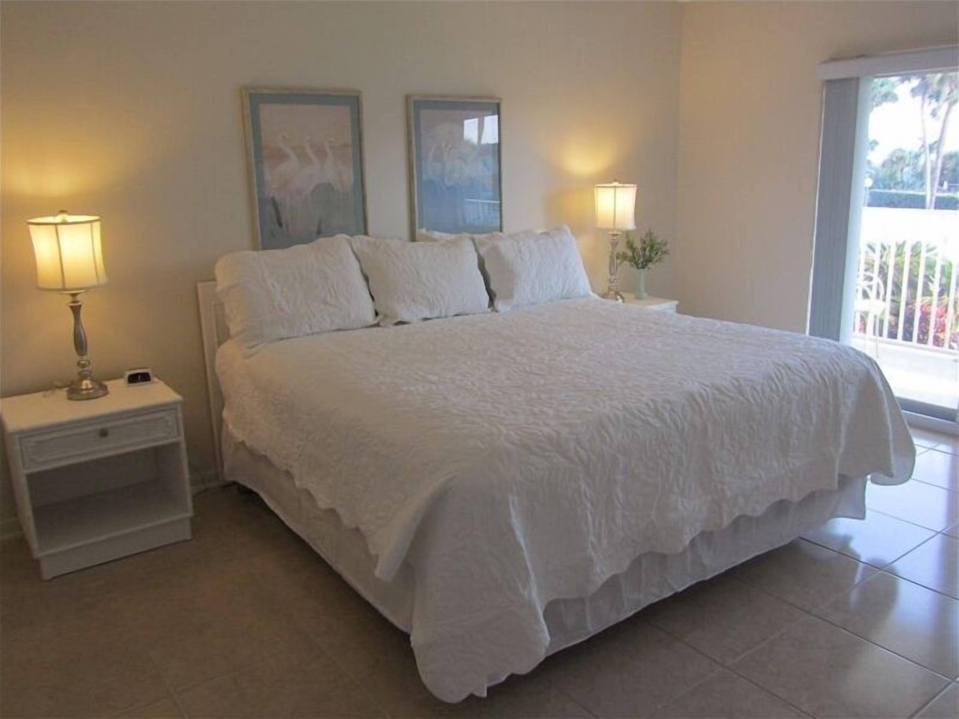 Una cama grande con ropa de cama blanca, dos mesitas de noche con lámparas y dos cuadros en la pared.