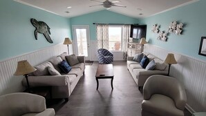 A living room with two sofas, a coffee table, and a television. There is a ceiling fan, a door leading to a balcony, and decorative wall art.