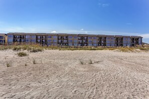 Ein mehrstöckiges Hotel mit flachem Dach und großen Fenstern, gelegen an einem Sandstrand mit spärlicher Vegetation.