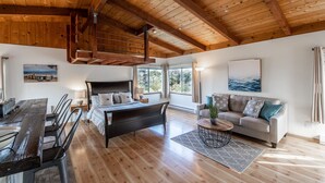A spacious room with a wooden ceiling, a bed, a sofa, a dining table, and a large window providing a view of the outdoors. - Stunning Oceanview Coastal Home – Beach, Trails, Family Activities (Montara)