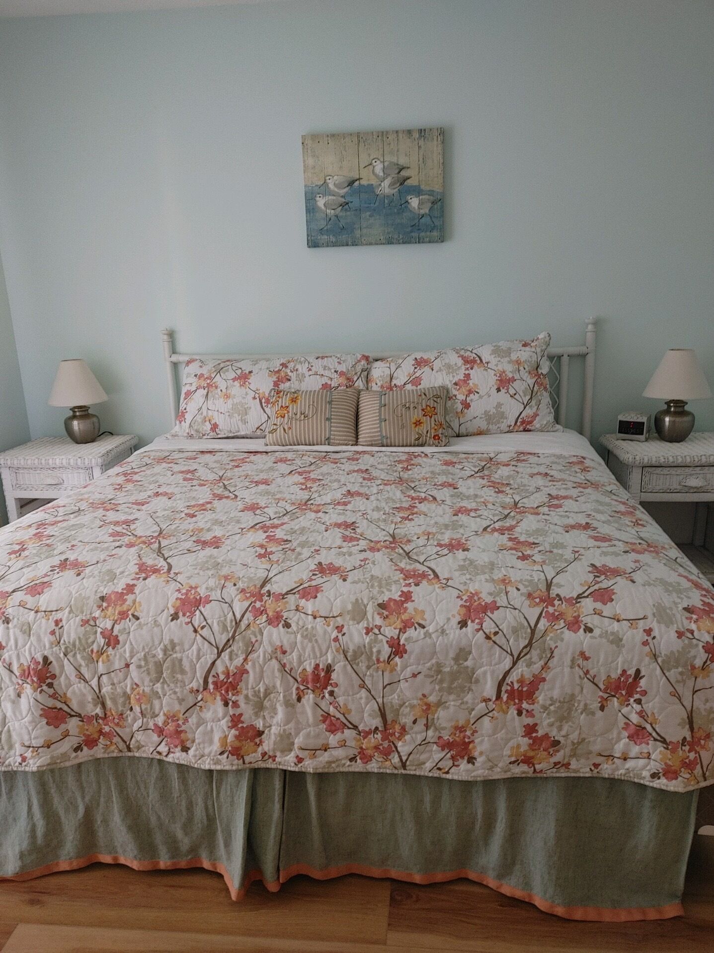 Een slaapkamer met een bloemenbedekking, twee nachtkastjes met lampen, een wanddecoratie en een lichtblauwe muur.
