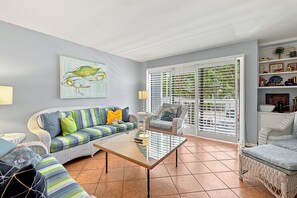A living room with a striped sofa, a glass coffee table, wicker chairs, and a large window with blinds.