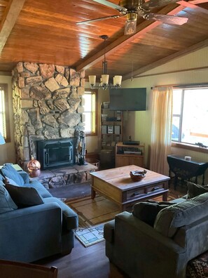 A cozy living room with a stone fireplace, wooden ceiling, and a flat-screen TV mounted on the wall. - Rent for Labor Day! Magnificent View! By golf course! Rates are very affordable! (Cloudcroft)