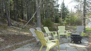 Eine Steinterrasse mit Adirondack-Stühlen und einer Feuerstelle in bewaldeter Landschaft.