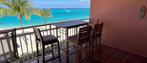 A balcony with a table and chairs overlooking a beach with clear blue water and a palm tree.