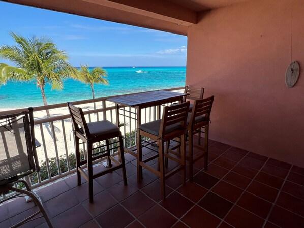 A balcony with a table and chairs overlooking a beach with clear blue water and a palm tree.