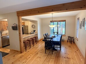 Uma sala de jantar com mesa e cadeiras de madeira, cozinha ao fundo e uma janela grande com persianas.