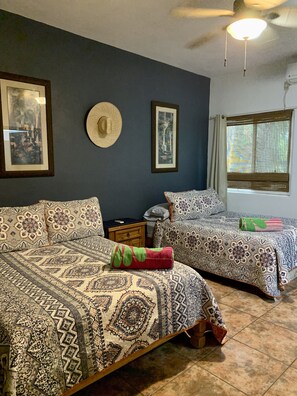 A bedroom with two beds, patterned bedding, a ceiling fan, framed artwork, and a window with a view.