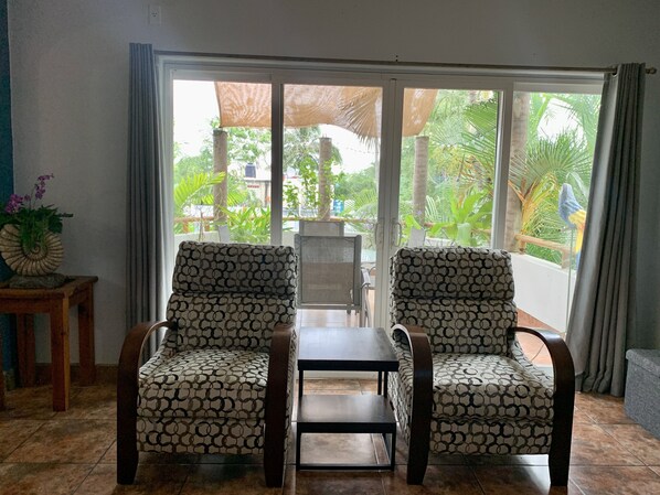 A room with two patterned armchairs, a small table, and a view of greenery through large sliding glass doors. - Villa Papaya, 2 bedrooms, specious, comfortable home, beach walkable. (San Francisco)