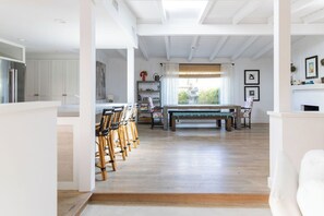 Um quarto amplo com piso de madeira, área de jantar com cadeiras e mesa, janela com cortinas e parede branca com quadros.