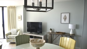Una sala de estar con televisión, una mesa de comedor, una silla, un sofá, una lámpara y un cuadro en la pared.