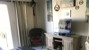 En stue med sofa, fjernsyn på et skab, stol og et vindue med gardiner.